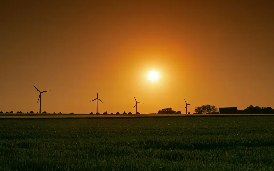 Silhouettes of wind turbines and a building against a bright orange sunset over a green field.