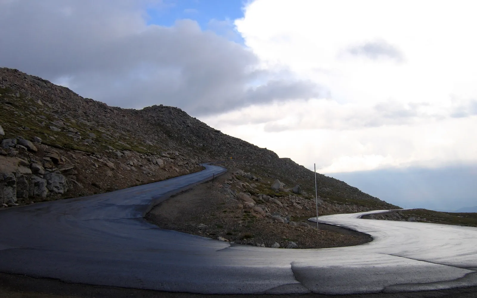 A winding asphalt road curves through a rocky, barren mountain landscape under a cloudy sky.
