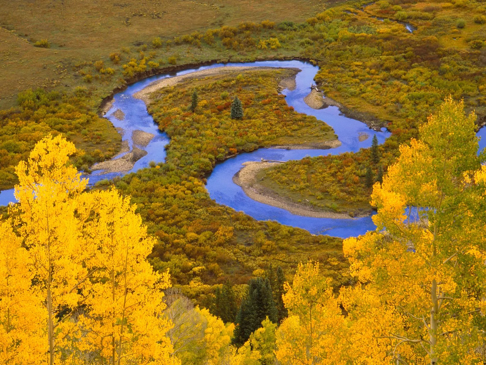 A bright blue river winds through a valley covered in golden and orange autumn foliage.
