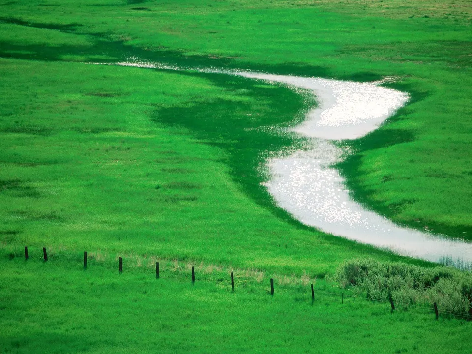 A bright, winding river flows through a vibrant green grassy landscape with a fence in the foreground.