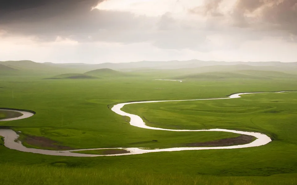 A wide, winding river flows through vast, rolling green grasslands under a dramatic, cloudy sky.