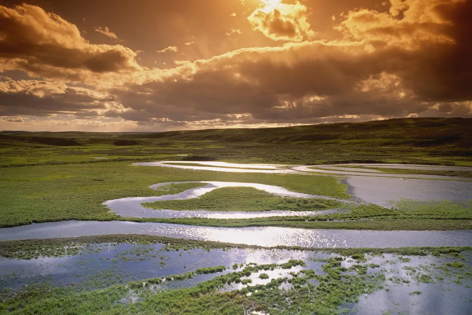 A wide, winding river flows through a green, grassy plain under a dramatic sunset sky.