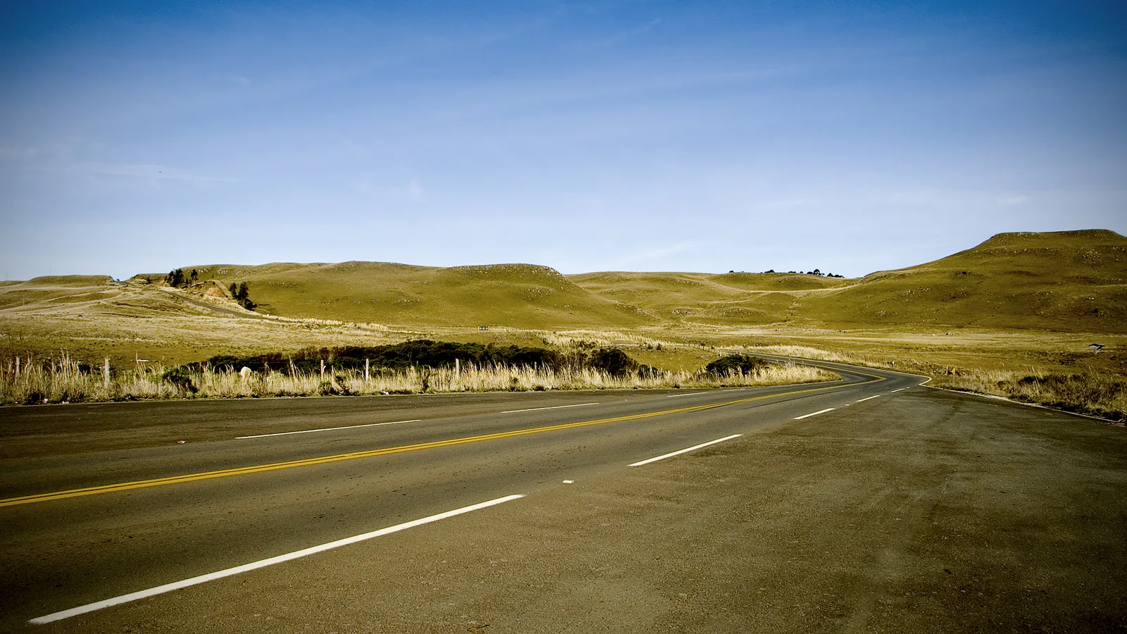 A paved road with yellow and white lines curves through a landscape of rolling, dry, golden hills under a clear blue sky.