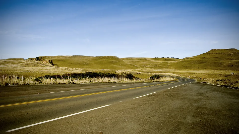 A paved road with yellow and white lines curves through a landscape of rolling, dry, golden hills under a clear blue sky.