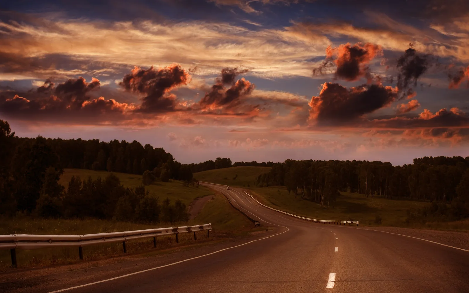 A winding road leads through rolling hills and forests under a dramatic sky filled with fiery clouds at sunset.