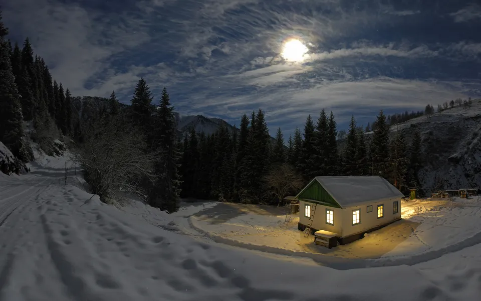 A small white cabin with lit windows sits in a snowy landscape under a bright moon and cloudy sky.
