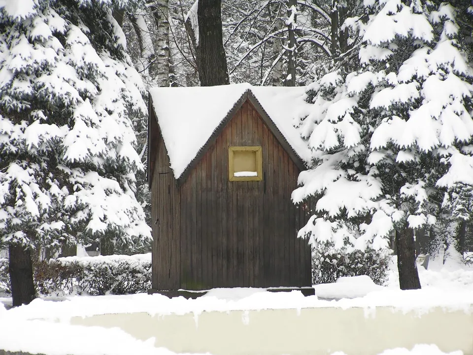 A small wooden cabin with a snow-covered roof sits amidst snow-laden evergreen trees.