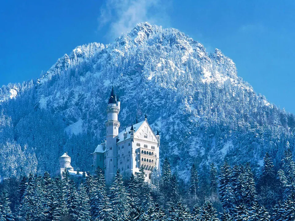 A majestic white castle sits nestled in a snow-covered forest at the base of a large, snow-capped mountain.
