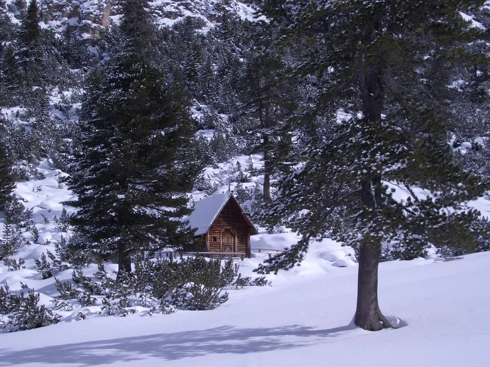 A small wooden chapel nestled in a snow-covered forest with pine trees.