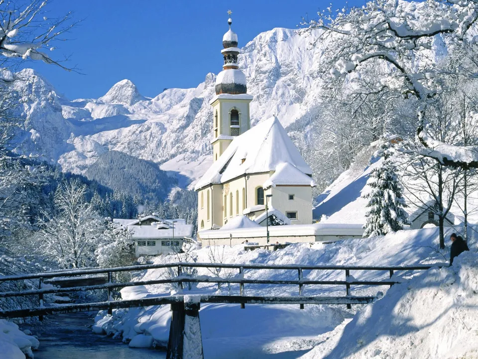 A snow-covered church stands in a winter landscape with majestic mountains behind it.