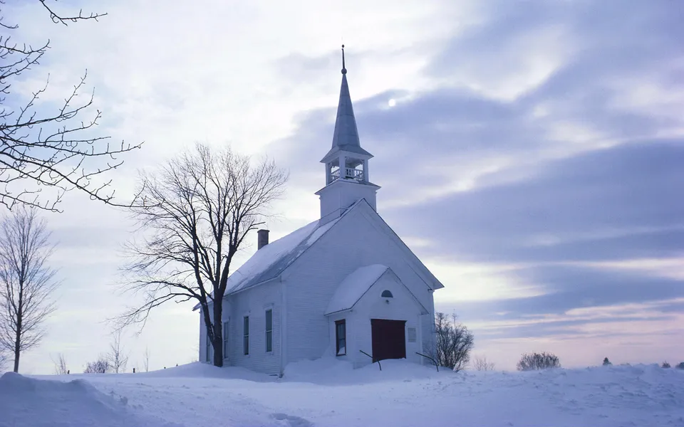 A white wooden church with a steeple sits in a landscape covered in deep snow under a cloudy sky.