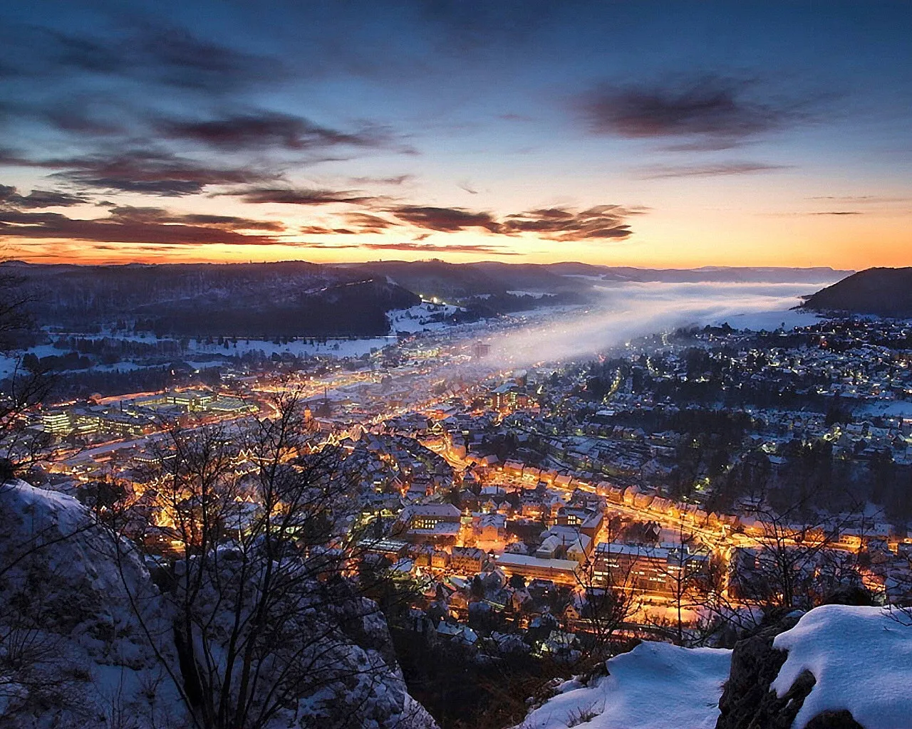 An aerial view of a snow-covered town at dusk, with lights twinkling and mist rising from a valley.