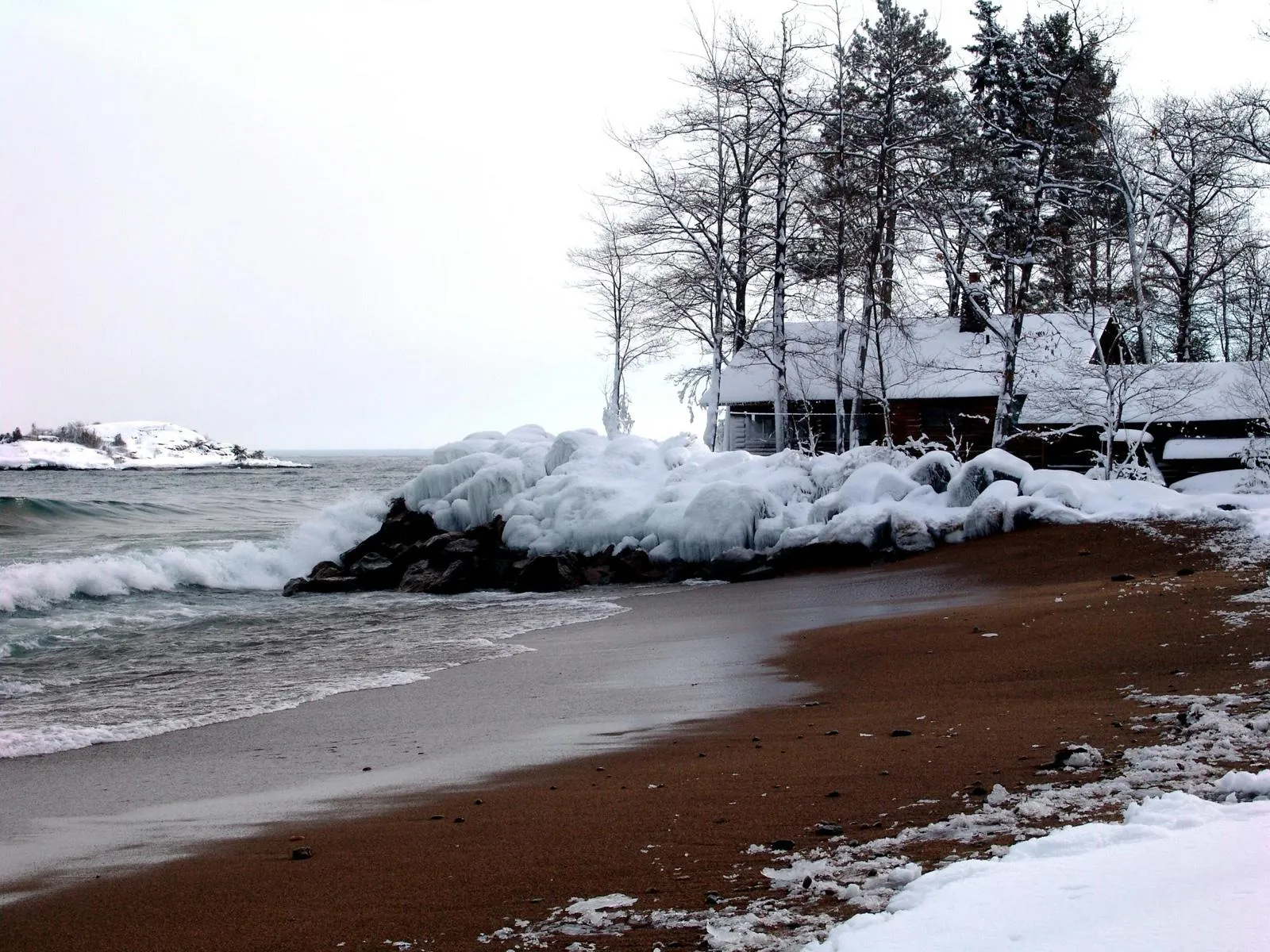 Waves crash on a sandy beach covered in snow and ice, with trees and cabins in the background.