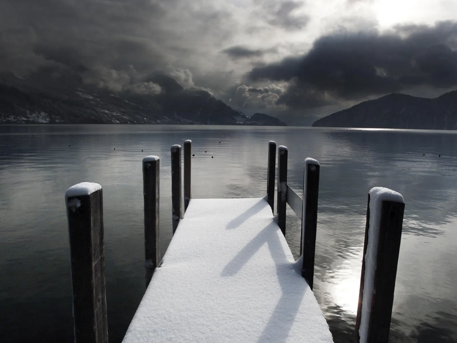 A wooden dock covered in snow extends into a calm lake, with mountains and dark clouds in the background.