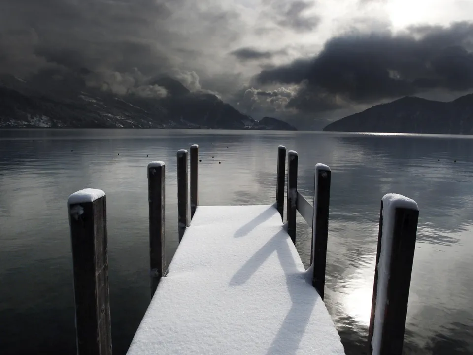 A wooden dock covered in snow extends into a calm lake, with mountains and dark clouds in the background.
