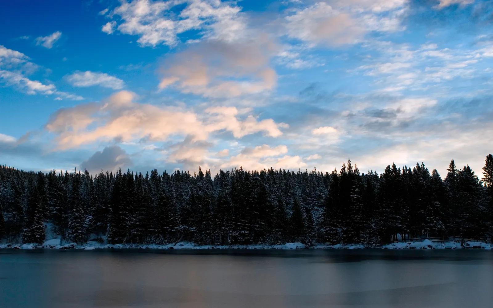 A dense forest of snow-covered pine trees borders a calm, partially frozen lake under a cloudy blue sky.