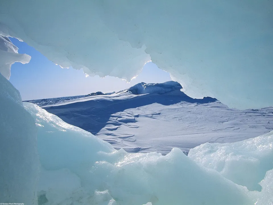 A view through a natural archway of ice and snow, revealing a vast, snow-covered landscape under a clear blue sky.