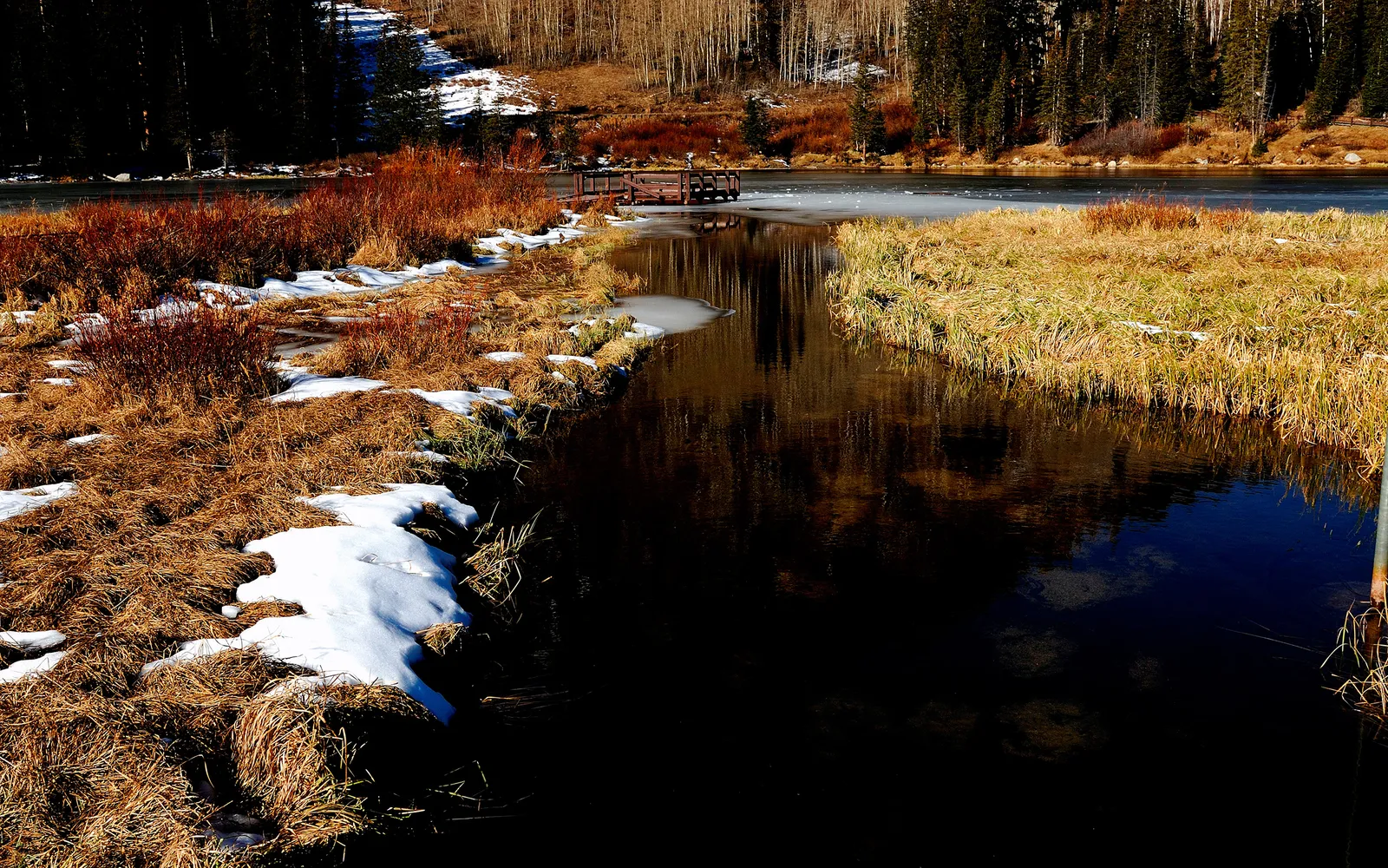 A frozen lake with a wooden dock, surrounded by dry grass and patches of snow.