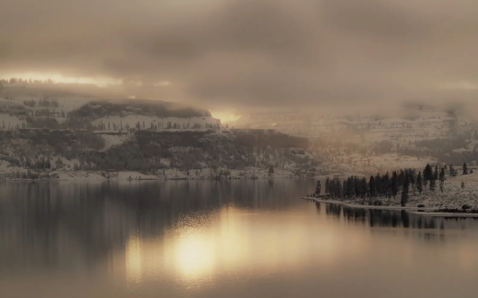 A serene winter lake reflects the muted light of a misty sunrise behind snow-covered hills.