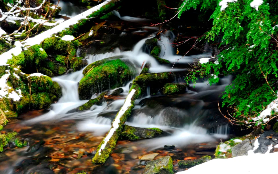 A long exposure shot of a stream with moss-covered rocks and snow.