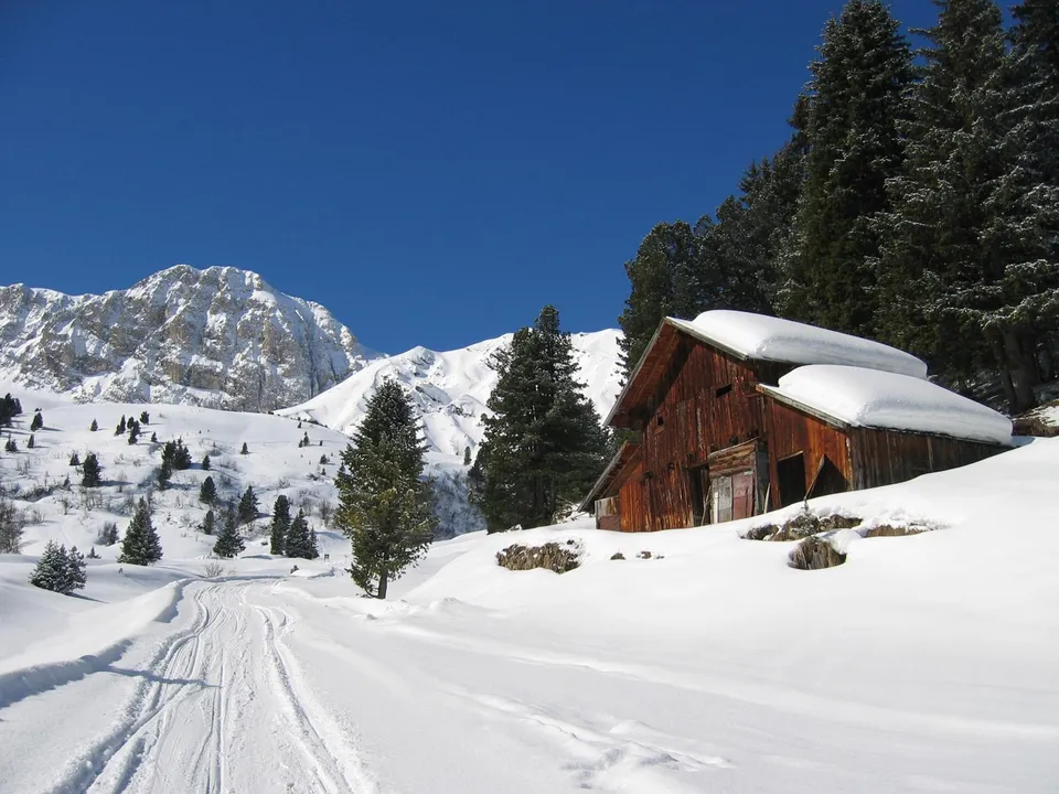 A wooden cabin sits in a snow-covered mountain landscape with tire tracks leading towards it.