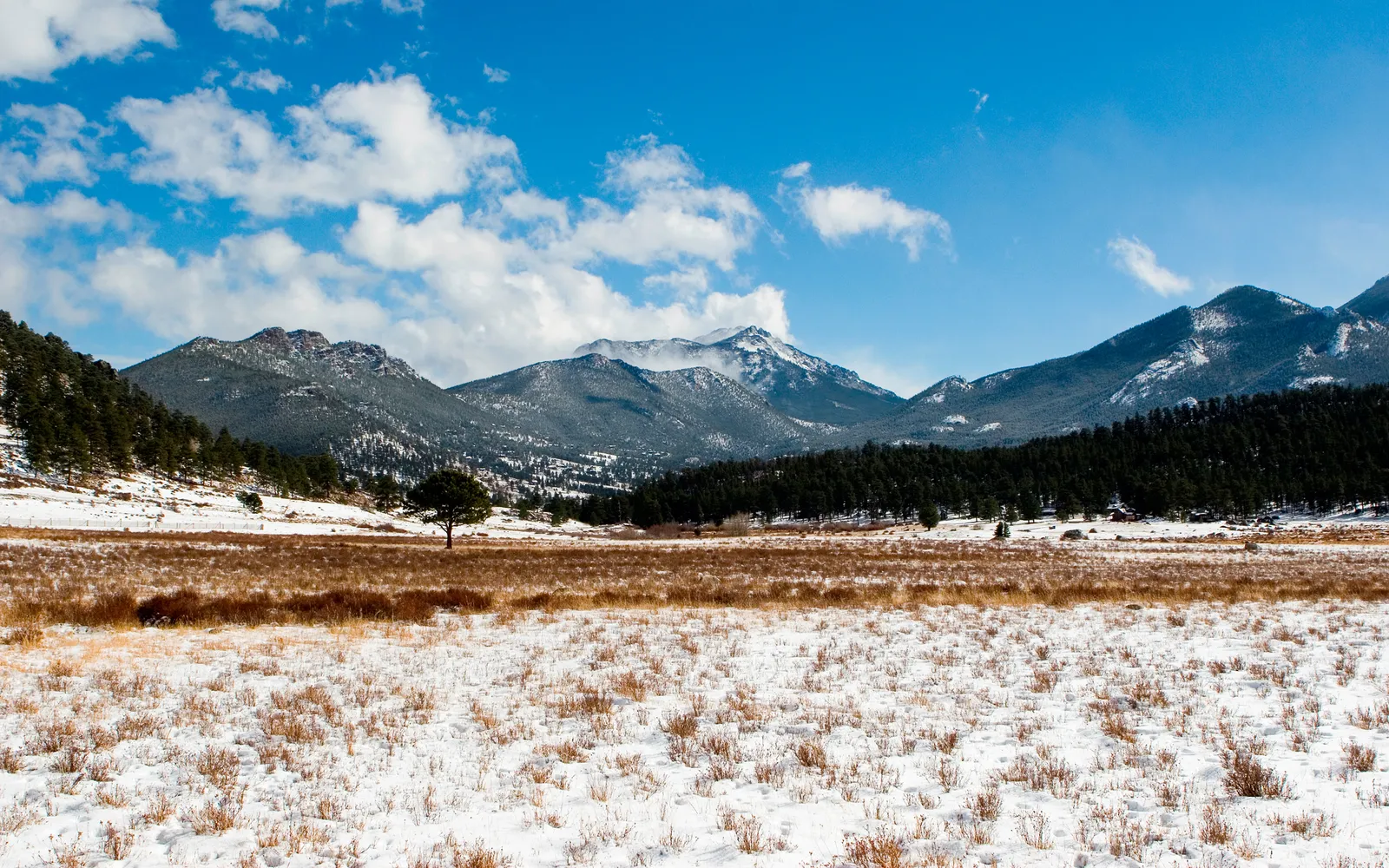 A wide view of snow-covered mountains and fields under a bright blue sky with scattered clouds.