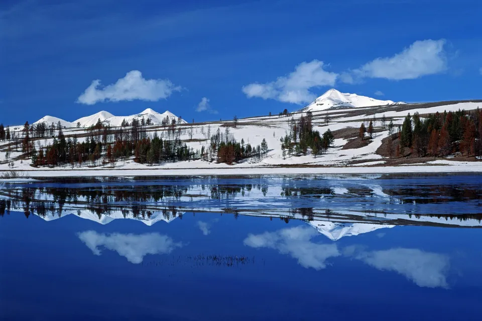 Snow-capped mountains and clouds are reflected in a deep blue lake under a bright sky.