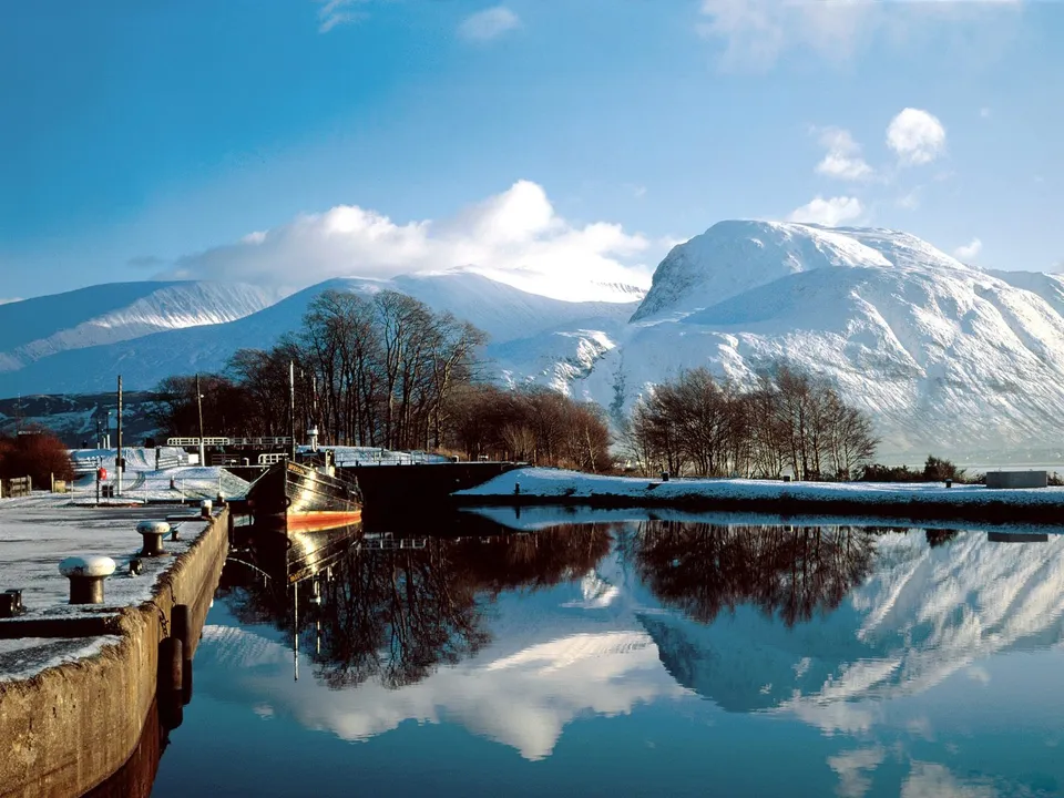 A canal with a boat and its reflection, leading to snow-covered mountains under a bright blue sky with clouds.