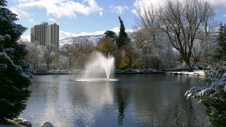 A park lake with a fountain in the center, surrounded by snow-covered trees and a tall building in the background.