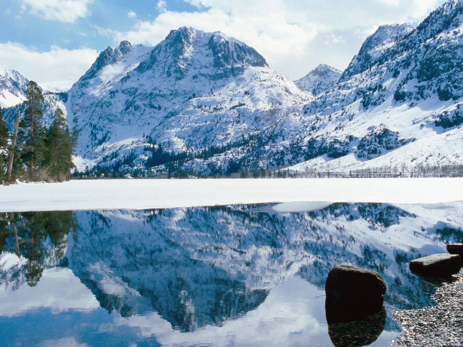 Snow-covered mountains are reflected in a partially frozen lake under a cloudy sky.