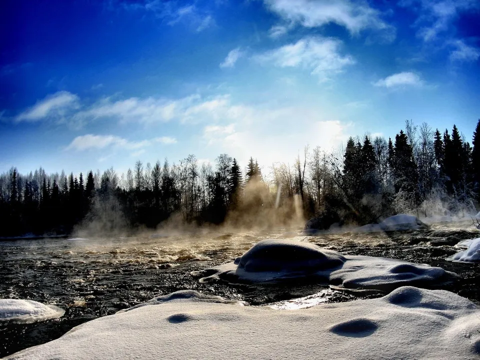 A misty river flows through a snow-covered landscape with a dense forest in the background under a bright blue sky.