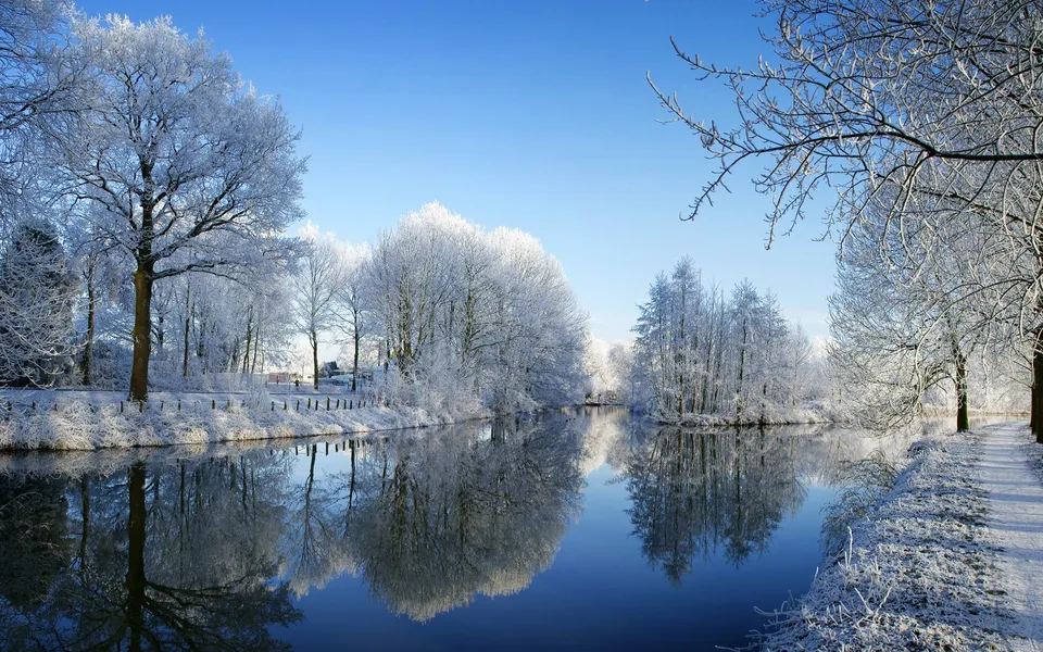 A calm river reflects snow-covered trees under a clear blue sky.