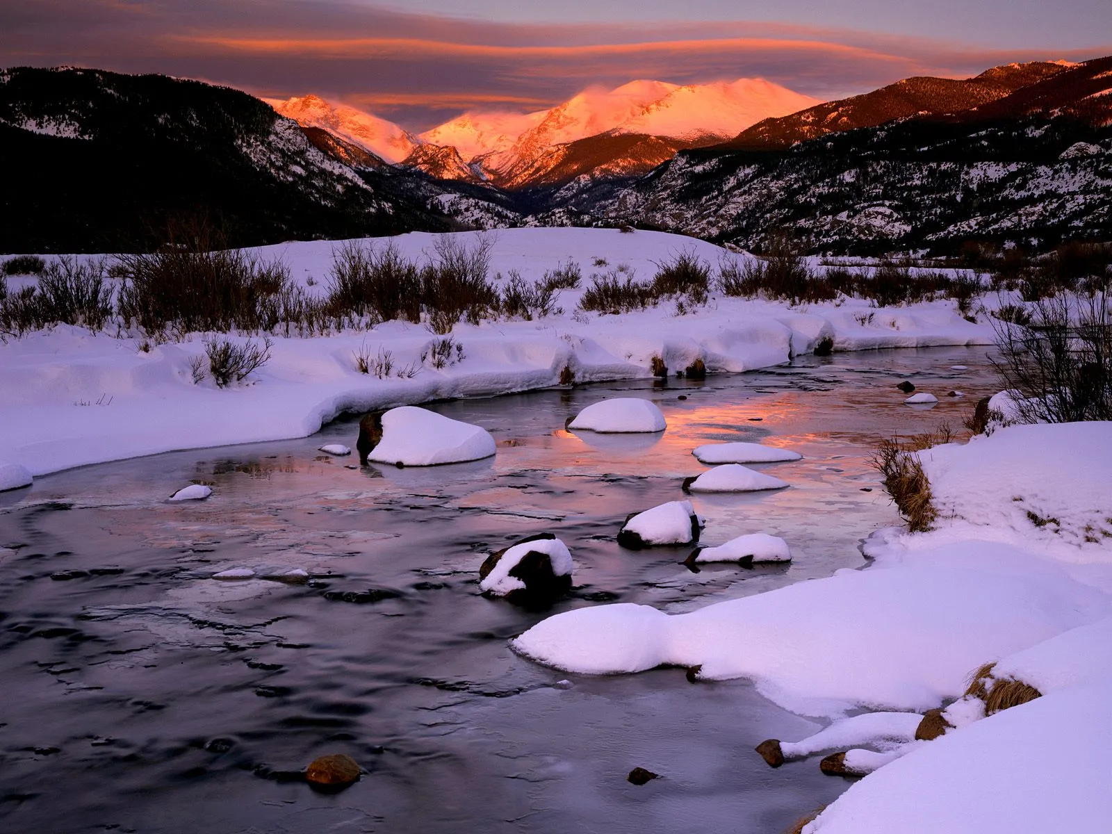A partially frozen river winds through a snow-covered valley, reflecting the sunset sky and distant snow-capped mountains.