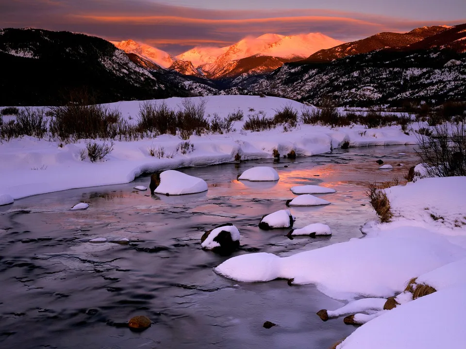A partially frozen river winds through a snow-covered valley, reflecting the sunset sky and distant snow-capped mountains.