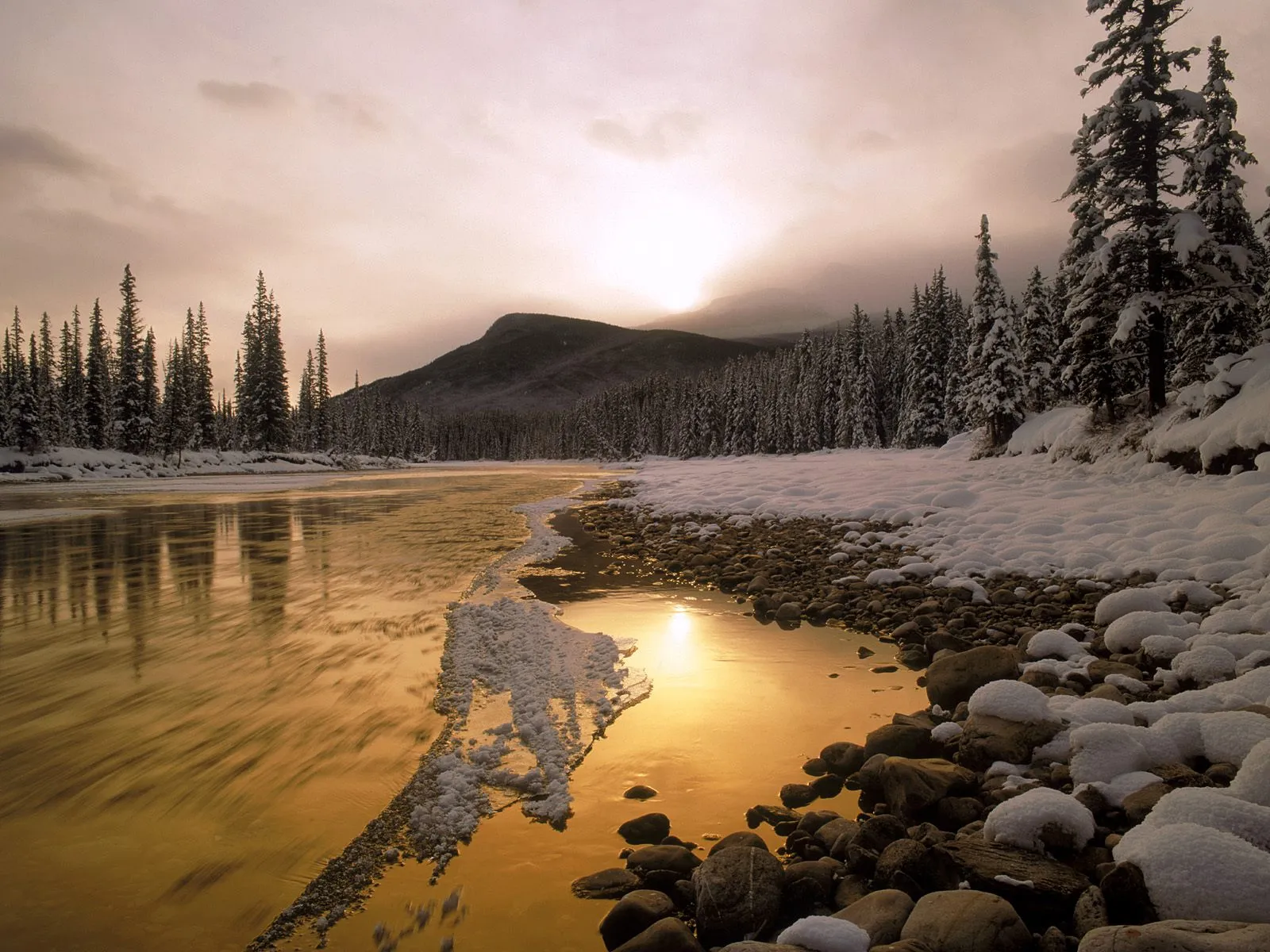 A golden sunset reflects on a partially frozen riverbank lined with snow-covered trees and rocks.
