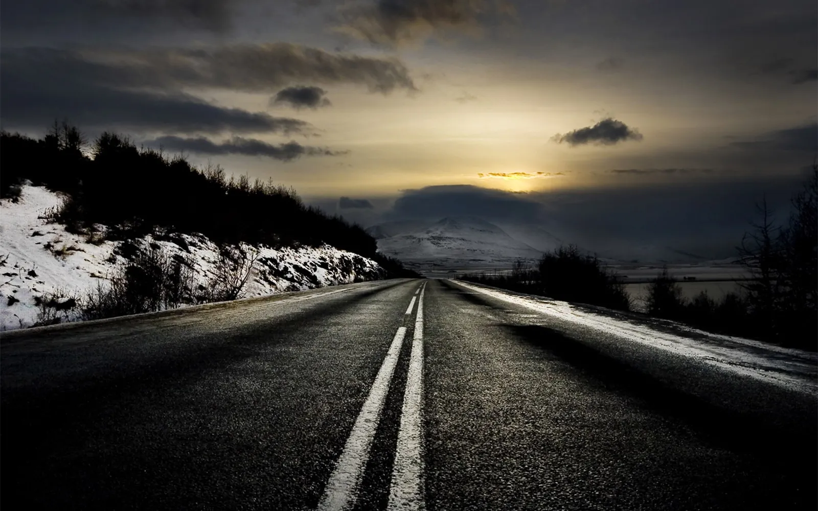 A dark, empty road winds through a snowy landscape under a dramatic, cloudy sky at dusk.
