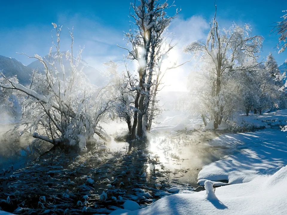 A sunlit winter scene with snow-covered trees and a partially frozen river emitting steam.