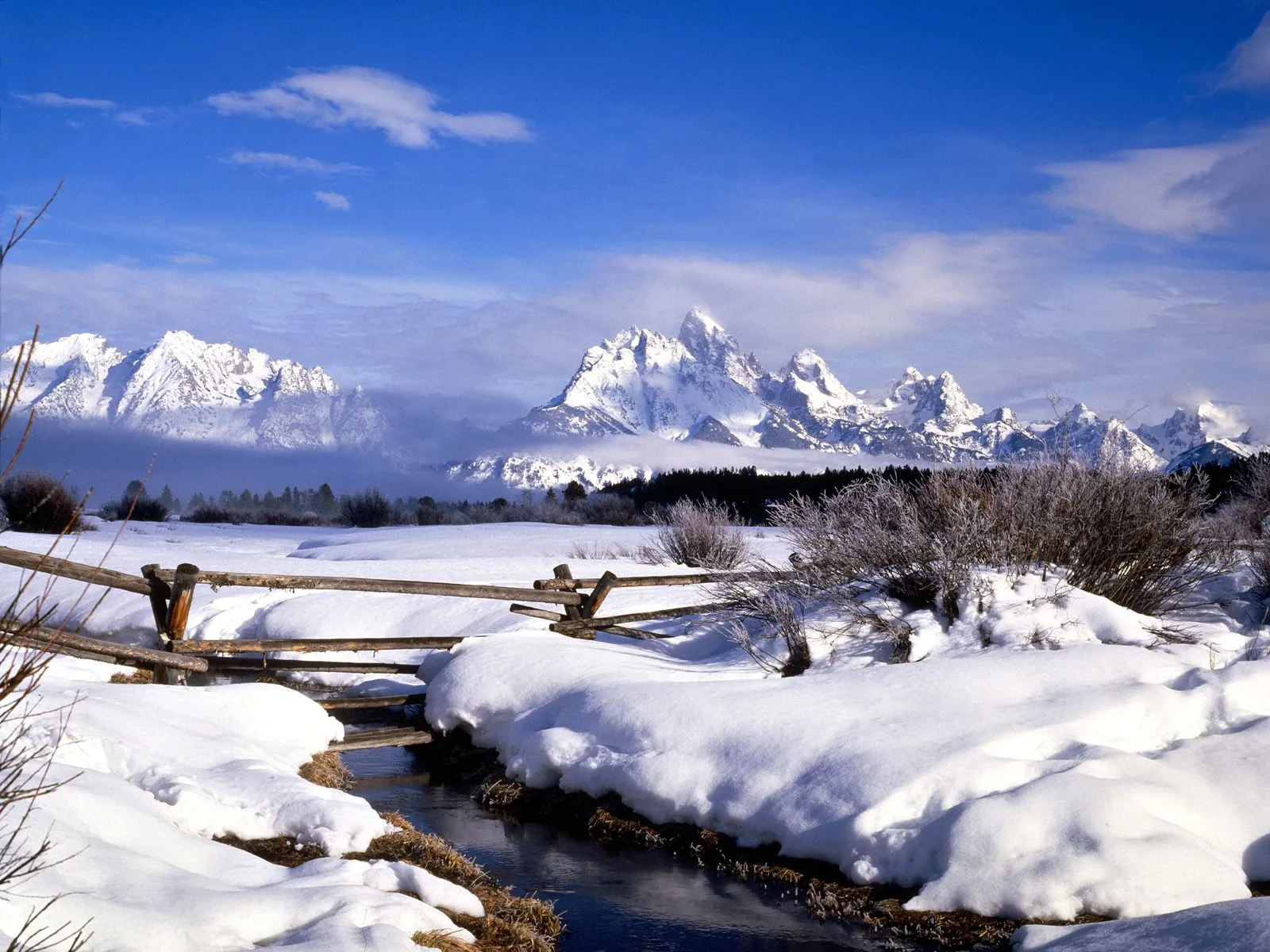 A snow-covered field with a small stream and a rustic wooden fence under a bright blue sky with scattered clouds.