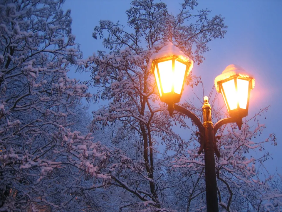 Two ornate street lamps glow warmly against a backdrop of snow-covered trees and a twilight sky.