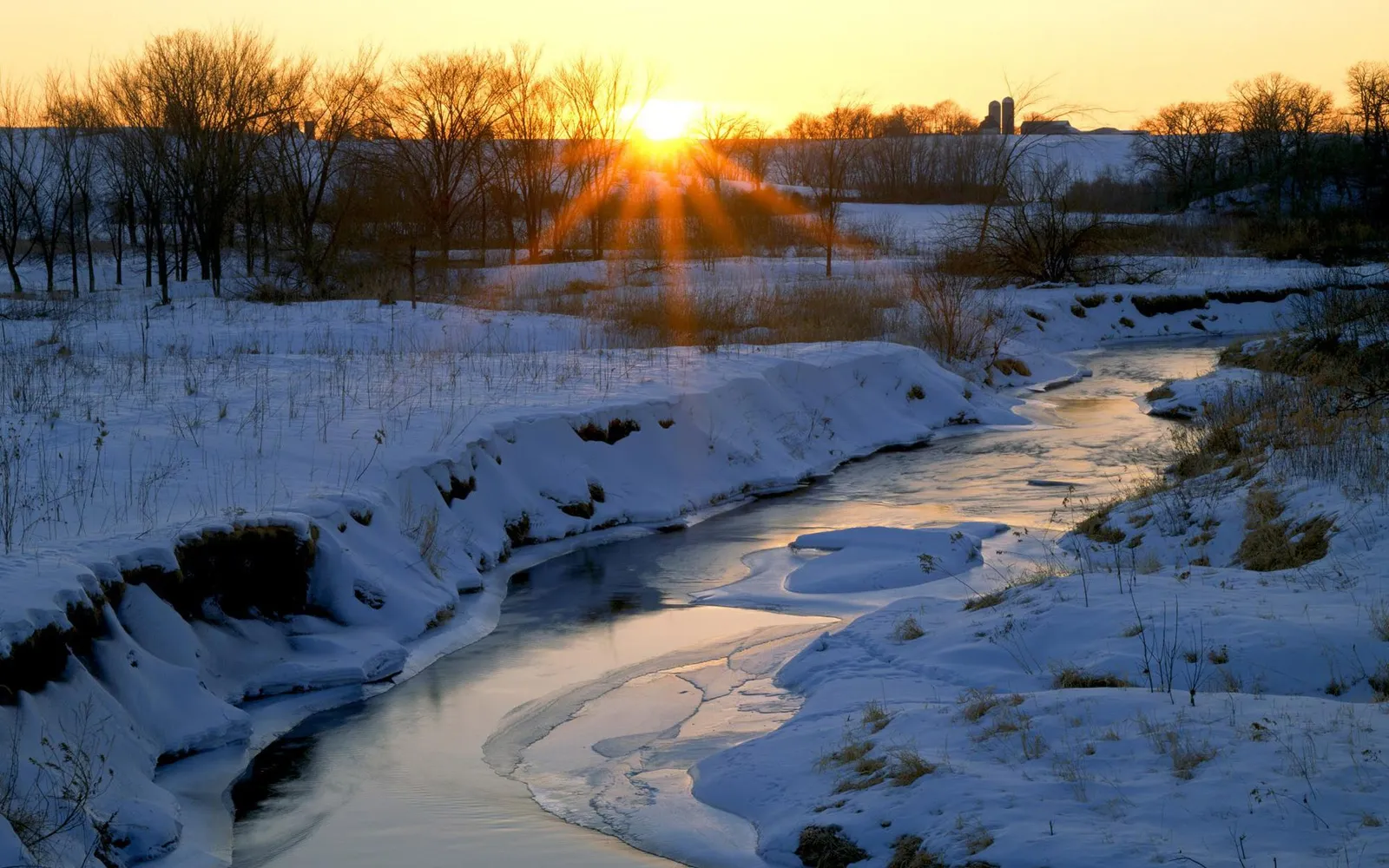 A winding river flows through a snow-covered landscape at sunrise, with the sun's rays bursting through bare trees.