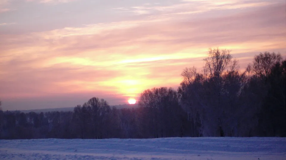 A vibrant sunset paints the sky with pink and yellow hues over a snow-covered landscape and silhouetted trees.