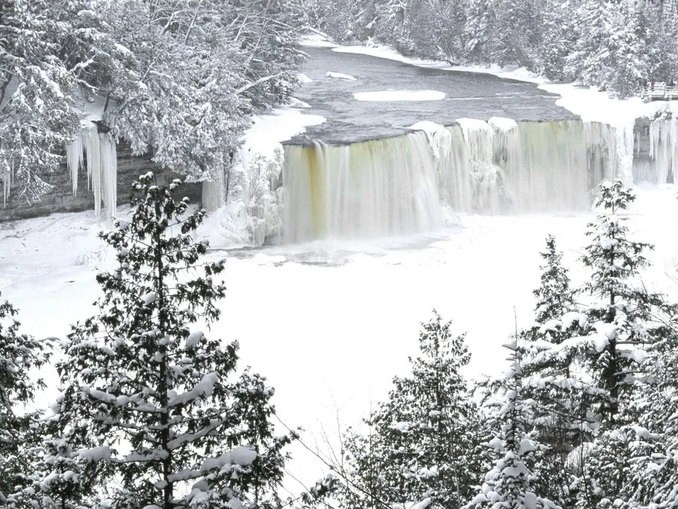 A wide waterfall cascades down a rocky cliff face, surrounded by snow-covered trees and icicles.
