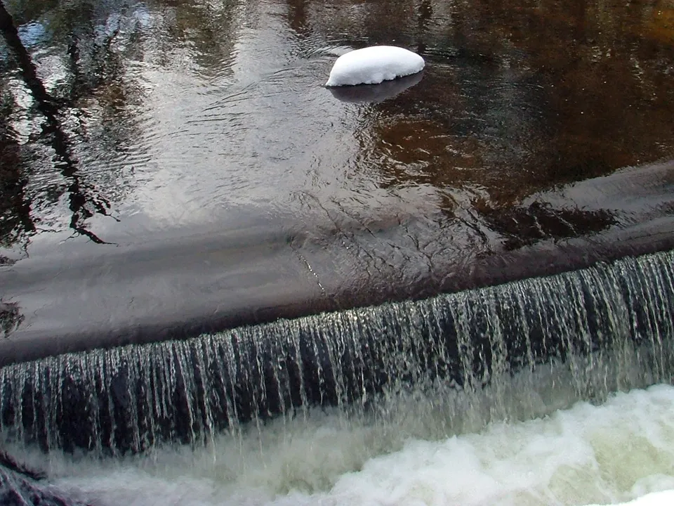 A small waterfall cascades over a dark rock ledge into foamy white water, with a patch of snow on the dark river surface.