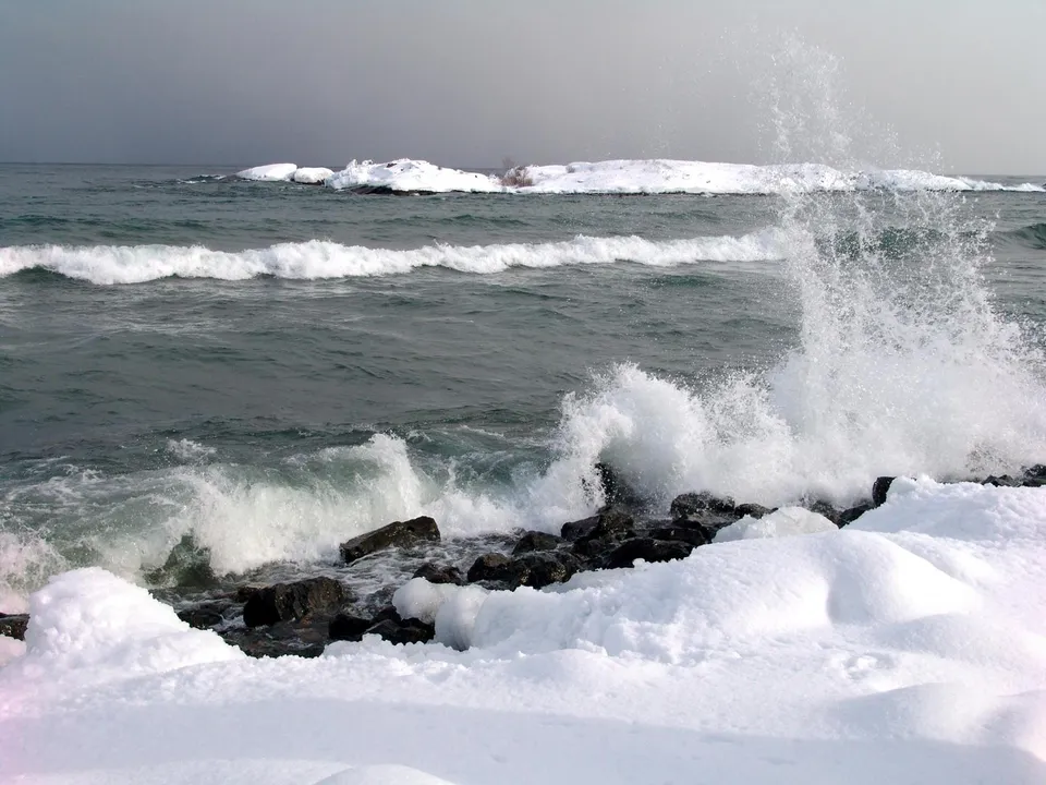 Waves crash against rocks and snow-covered shore with a distant island covered in snow.