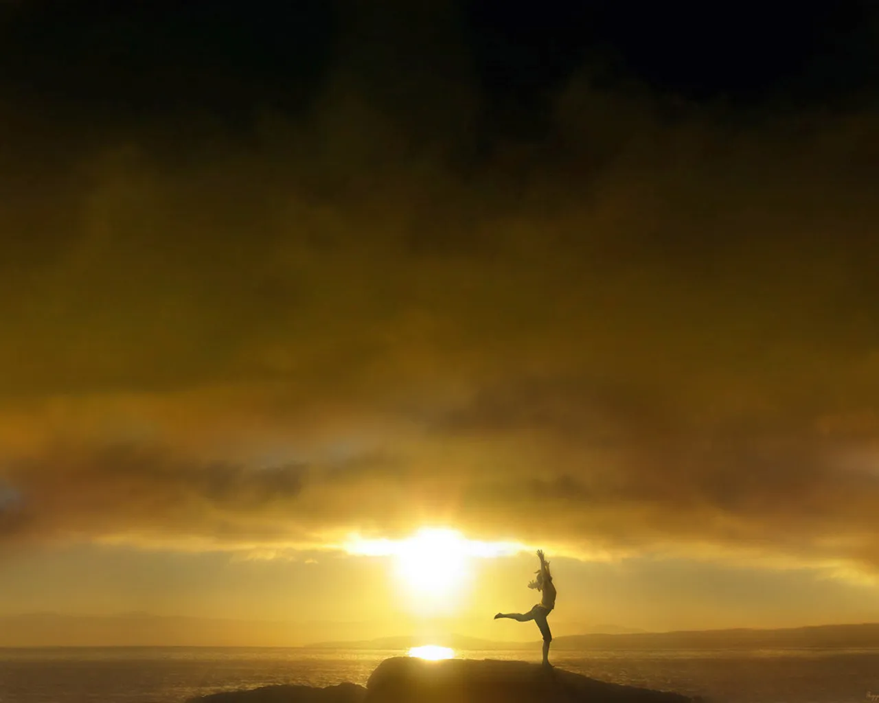 Silhouette of a woman in a yoga pose on a rock overlooking the ocean at sunset.