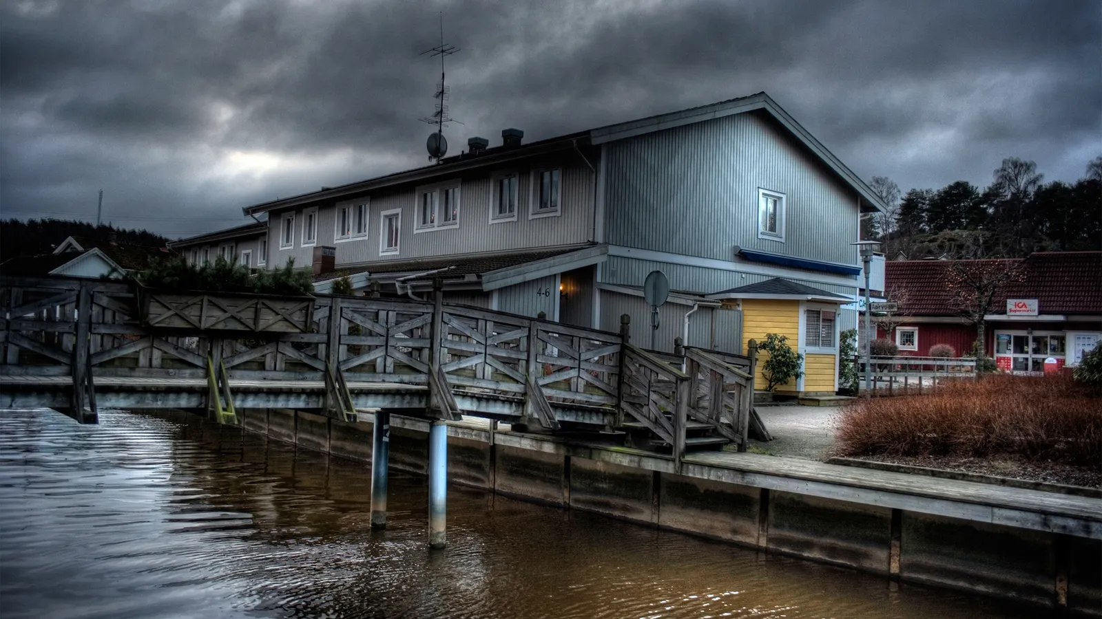 A wooden bridge crosses a calm body of water in front of buildings under a dark, cloudy sky.