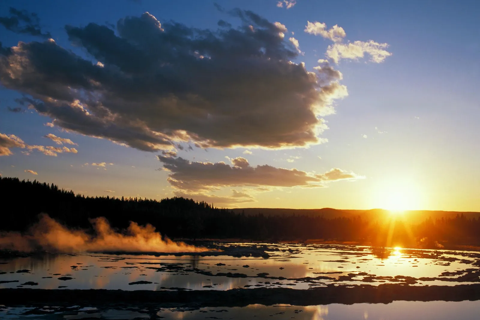A geothermal pool with steam rising, silhouetted trees, and a bright sunset over the horizon.