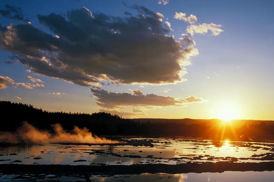 A geothermal pool with steam rising, silhouetted trees, and a bright sunset over the horizon.