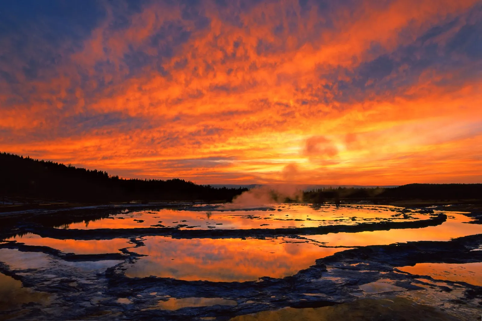 A dramatic sunset with fiery orange clouds reflects in the pools of a geothermal area with steam rising.