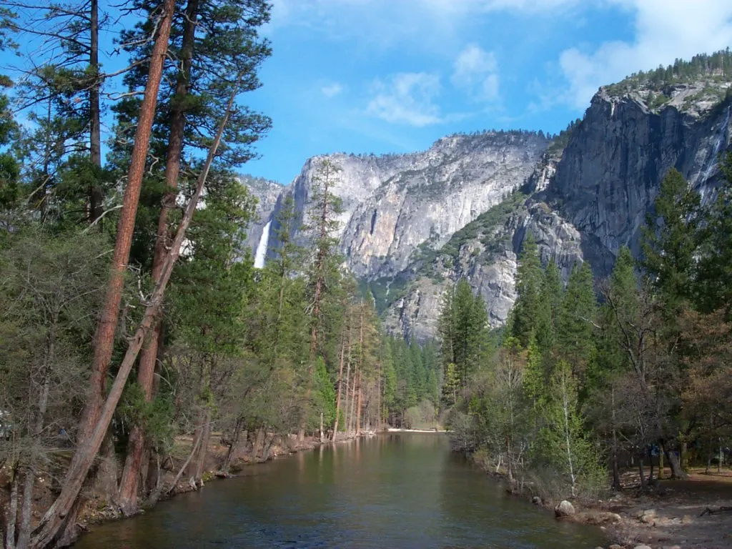 A wide river flows through a forest towards a tall waterfall cascading down a rocky mountain.
